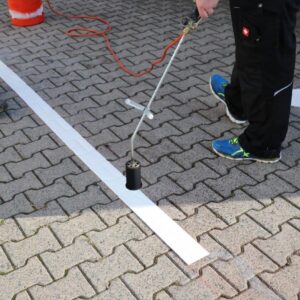 Arbeiter bringt mit einem Brenner weiße Bodenmarkierung auf Pflastersteinen an. bodenmarkierung-profishop.de