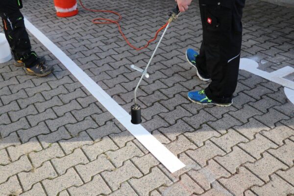 Arbeiter bringt mit einem Brenner weiße Bodenmarkierung auf Pflastersteinen an. bodenmarkierung-profishop.de